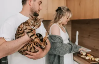 A man holding a Bengal cat while a woman slices fruits in a cozy kitchen setting.